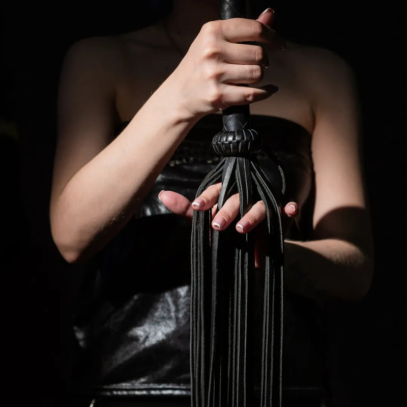 Person in black leather outfit holding a flogger whip - representing AdultSexToys.com’s bondage and BDSM collection.